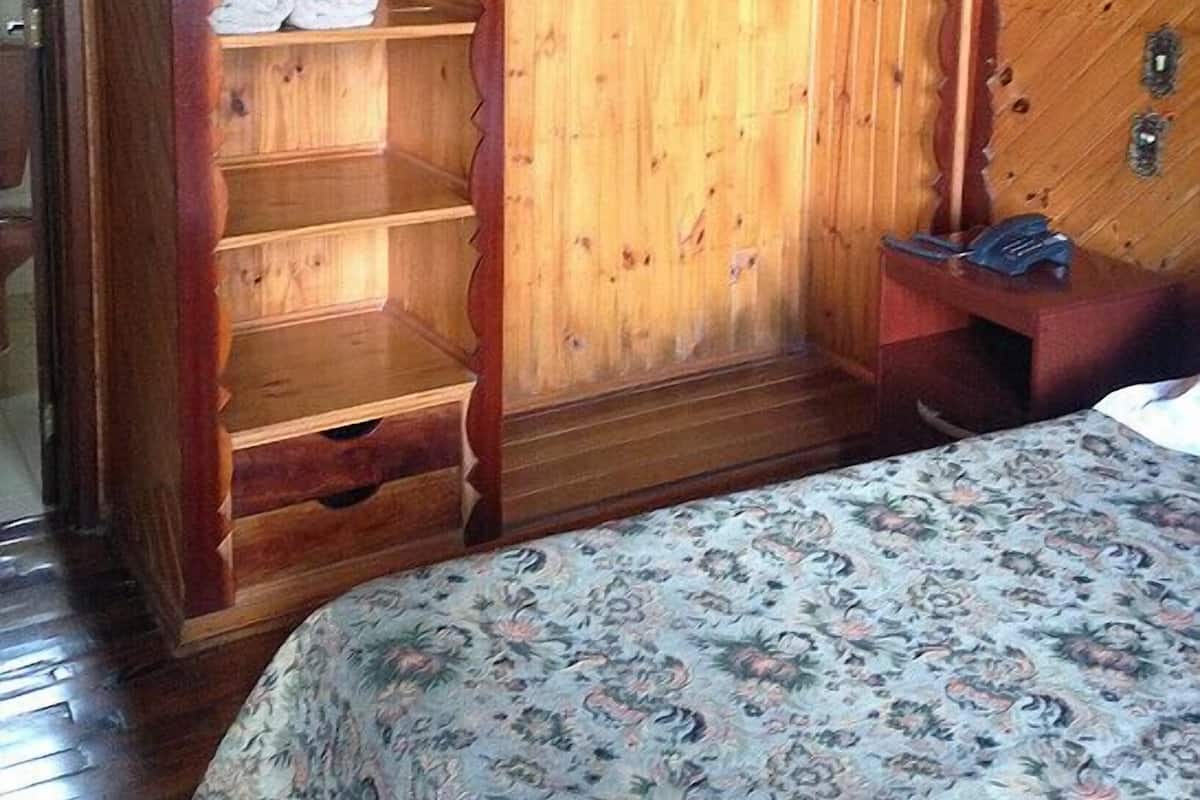 A wooden room with a sloped ceiling, a bed with a floral bedspread, and a wooden wardrobe with shelves.