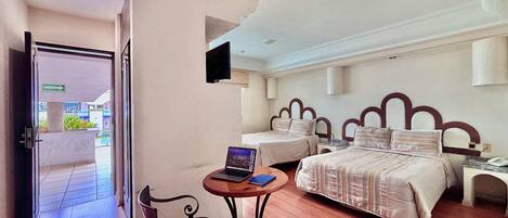 Quarto de hotel com duas camas, uma escrivaninha com um laptop, uma cadeira e uma televisão na parede.