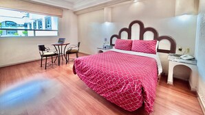 Habitación de hotel con cama, escritorio con computadora portátil, silla y ventana con vista a edificios.