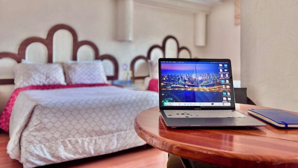 Una habitación de hotel con una cama, un ordenador portátil sobre una mesa de madera y un paisaje urbano en la pantalla del portátil.