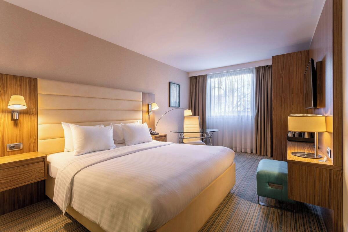 Ein Hotelzimmer mit einem großen Bett, Nachttischen mit Lampen, einem kleinen Schreibtisch mit Stuhl, einem an der Wand montierten Fernseher und einem Fenster mit Vorhängen.