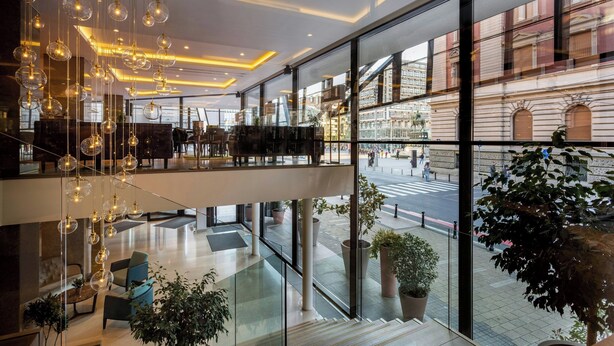 Een moderne hotellobby met glazen wanden, een entresol, hangende sferische lampen en potplanten.