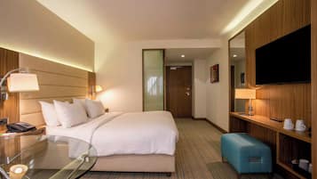 Quarto de hotel com cama, mesa de cabeceira, luminária, televisão e um pequeno espaço de estar.