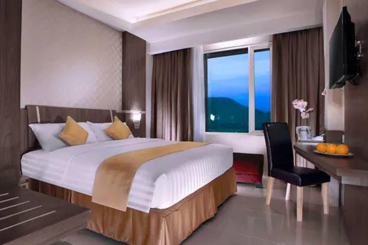 Habitación de hotel con una cama grande, un escritorio con silla, televisión y ventana con vista a la ciudad al atardecer.