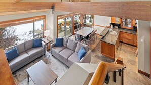 A spacious living area with a large sofa, a coffee table, and a dining table. There is a kitchen area with wooden cabinets and a window overlooking the outdoors. - Montebello by Whistler Premier (Whistler)
