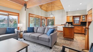 A spacious living area with a gray sofa, wooden ceiling beams, large windows, and a kitchen with wooden cabinets. - Montebello by Whistler Premier (Whistler)