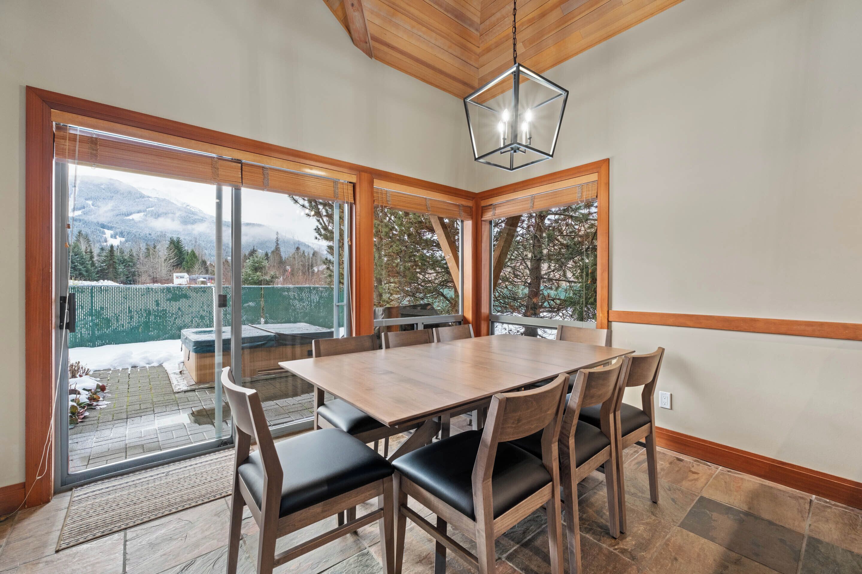 A dining area with a wooden table and chairs, large windows with a view of mountains and trees, and a hanging light fixture.