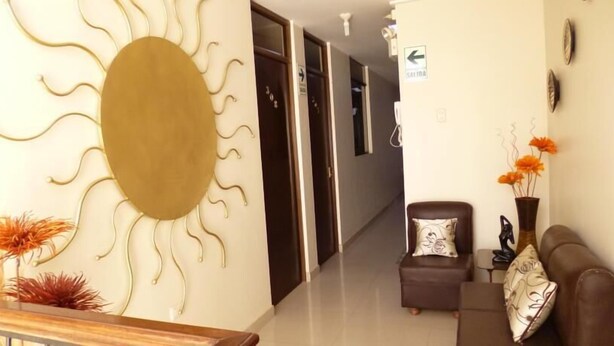 Un vestíbulo de hotel con un adorno de pared con forma de sol, un sofá con cojín floral y un jarrón con flores naranjas.