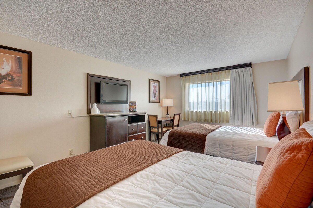 Una habitación de hotel con dos camas, un escritorio con silla, televisión y ventana con cortinas.