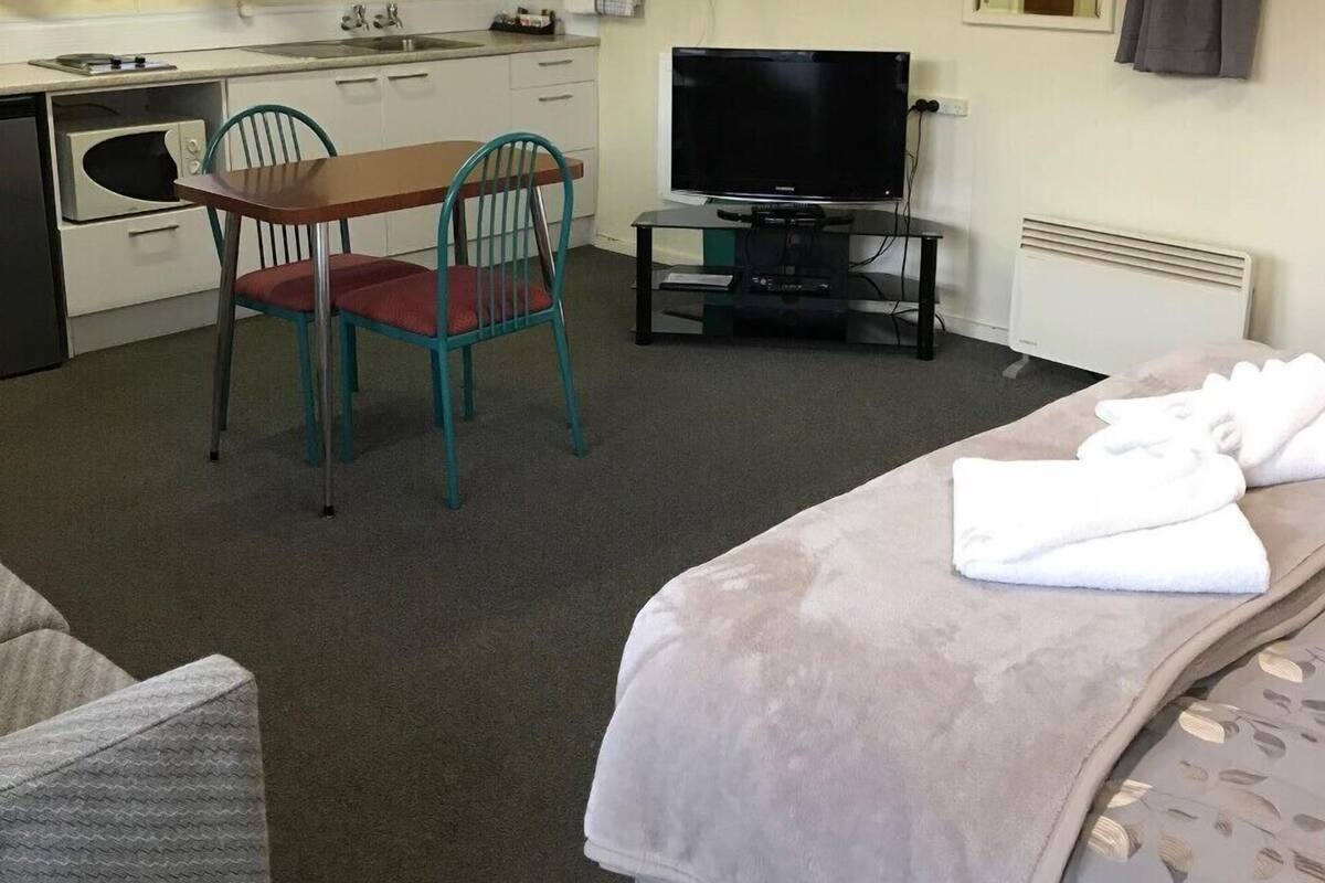 Ein kompaktes Hotelzimmer mit Bett, einer kleinen Küchenzeile, einem Esstisch mit Stühlen, einem Fernseher und einem Spiegel.