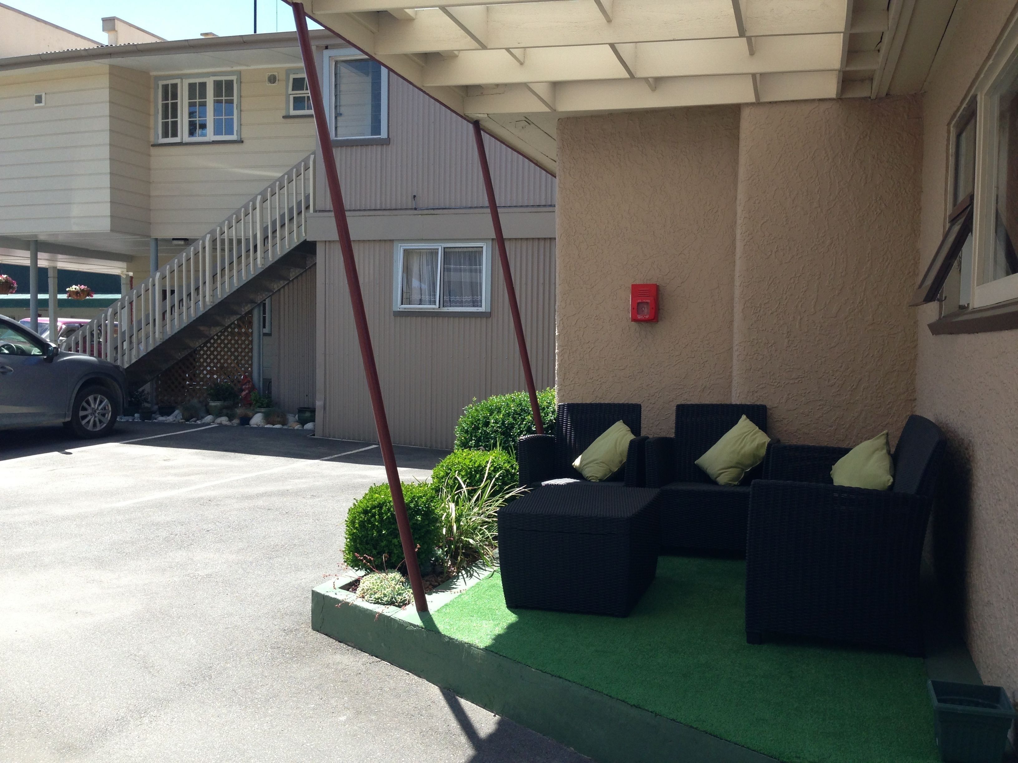 A covered outdoor seating area with a sofa and chairs, a staircase leading to a building, and a parked car.