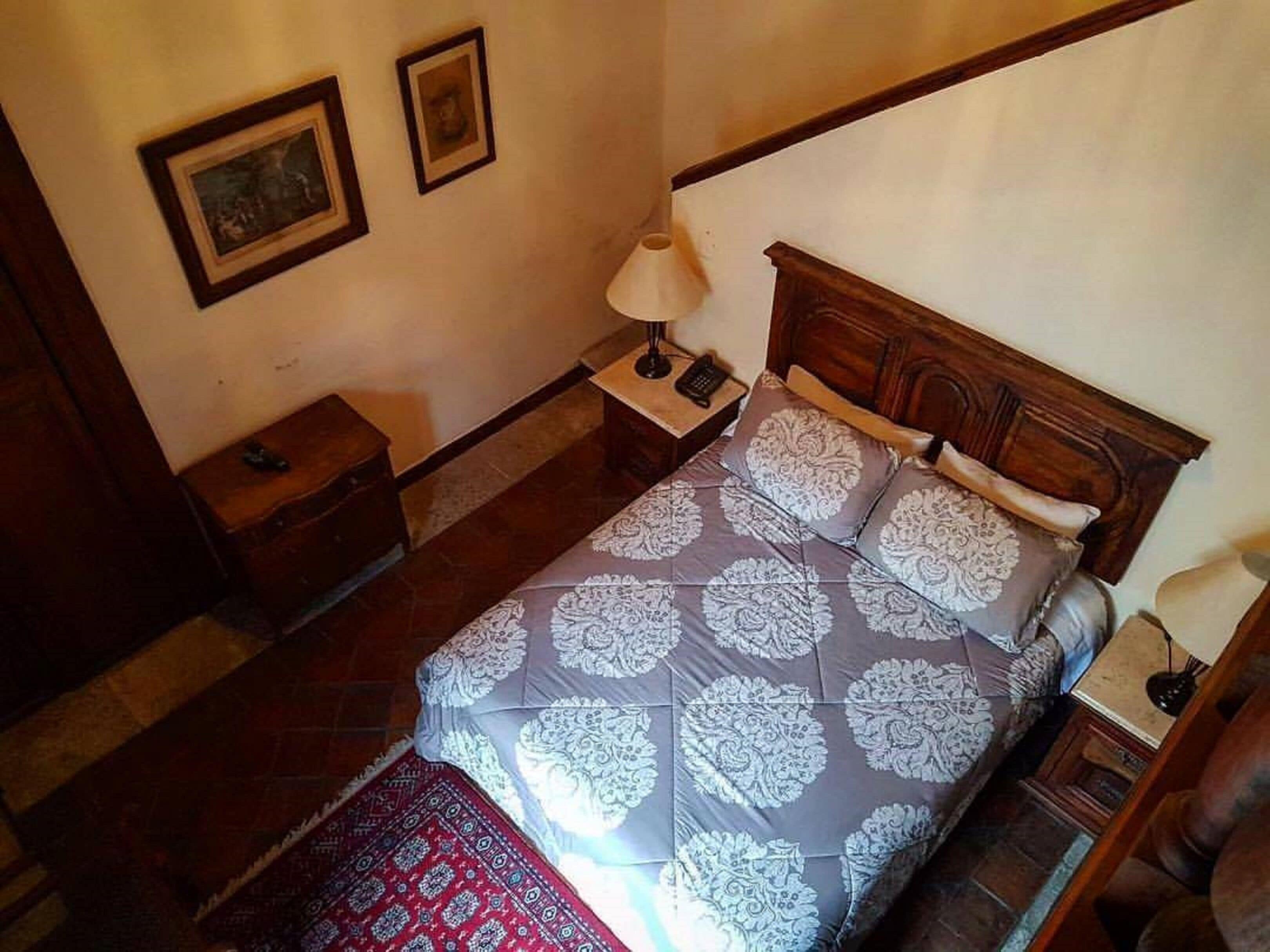 Un dormitorio con cabecera de madera, una cama con ropa de cama estampada, una mesita de noche con una lámpara y una pequeña mesa con un teléfono.