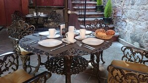 Une table à manger dressée pour quatre personnes, avec des chaises en métal décorées, un panier de pain, et un escalier en arrière-plan.