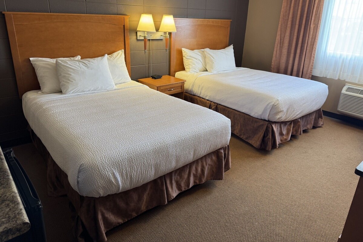 A hotel room with two beds, a headboard, bedside lamps, a nightstand, and a window with curtains.