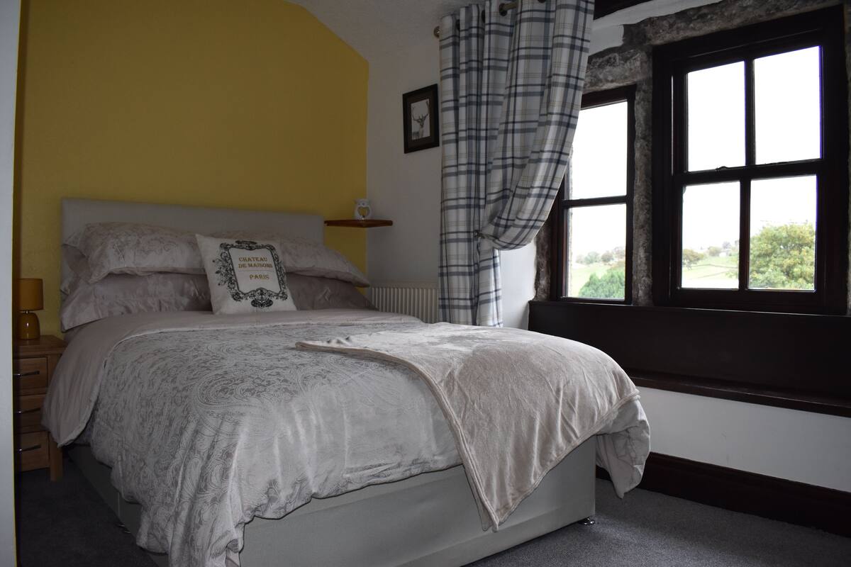 A bedroom with a bed, a window with curtains, a bedside table, and a wall-mounted shelf.
