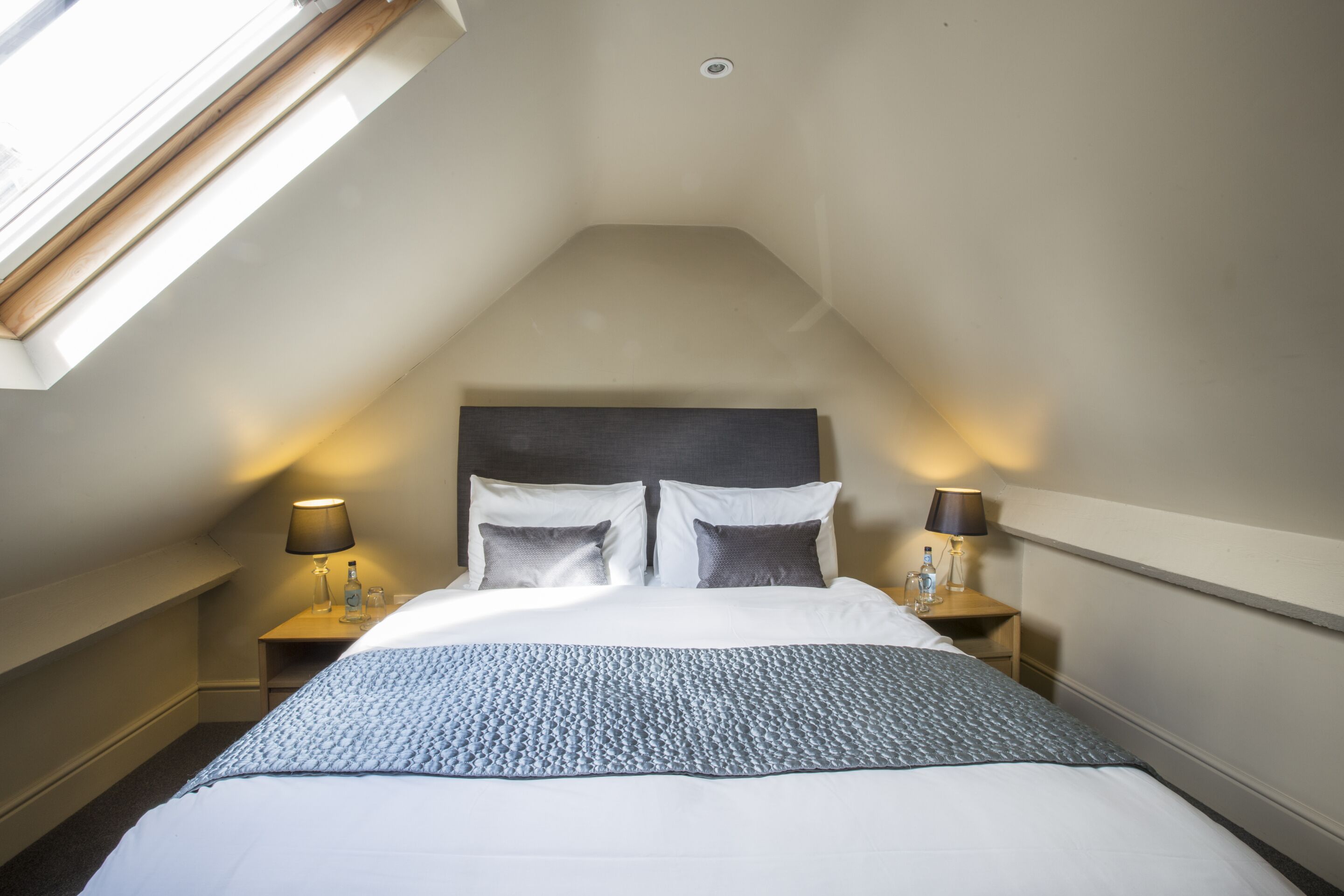 A bedroom with a sloped ceiling, a bed with white bedding, two bedside tables with lamps, and a skylight.