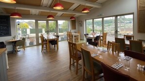 A spacious dining area with wooden tables and chairs, large windows offering a view of the outdoors, and red pendant lighting.