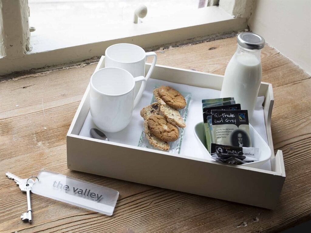 A tray with white mugs, cookies, a milk bottle, and a keychain labeled "the valley" on a wooden surface near a window.