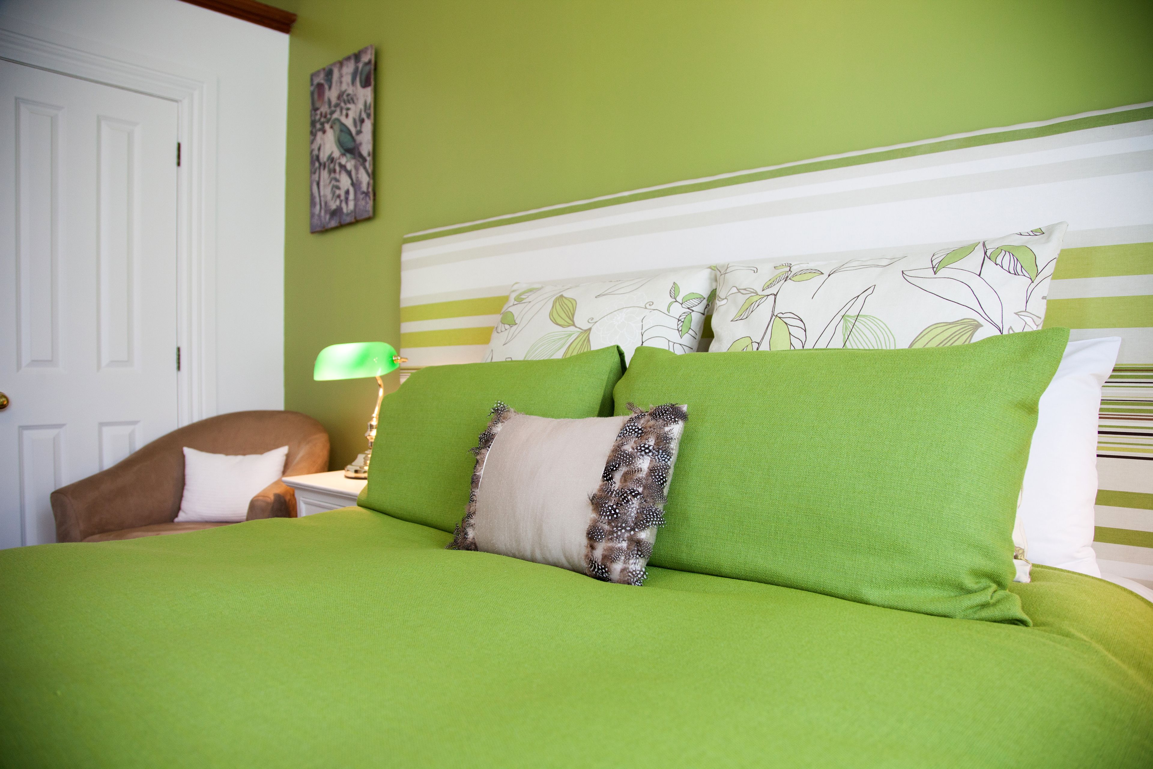 A bed with green bedding and a white headboard, a brown chair, a nightstand with a lamp, and a wall with a floral painting.