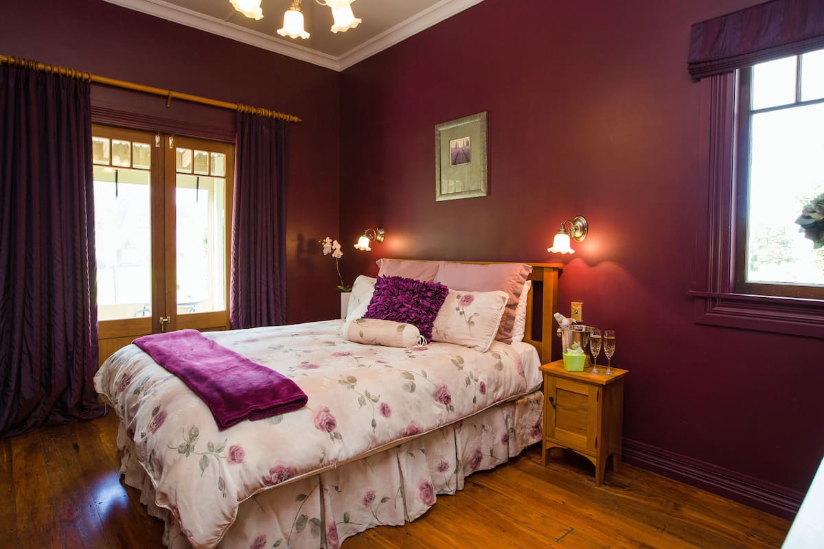 A bedroom with a floral bedspread, a wooden nightstand with two glasses, and a window with purple curtains.