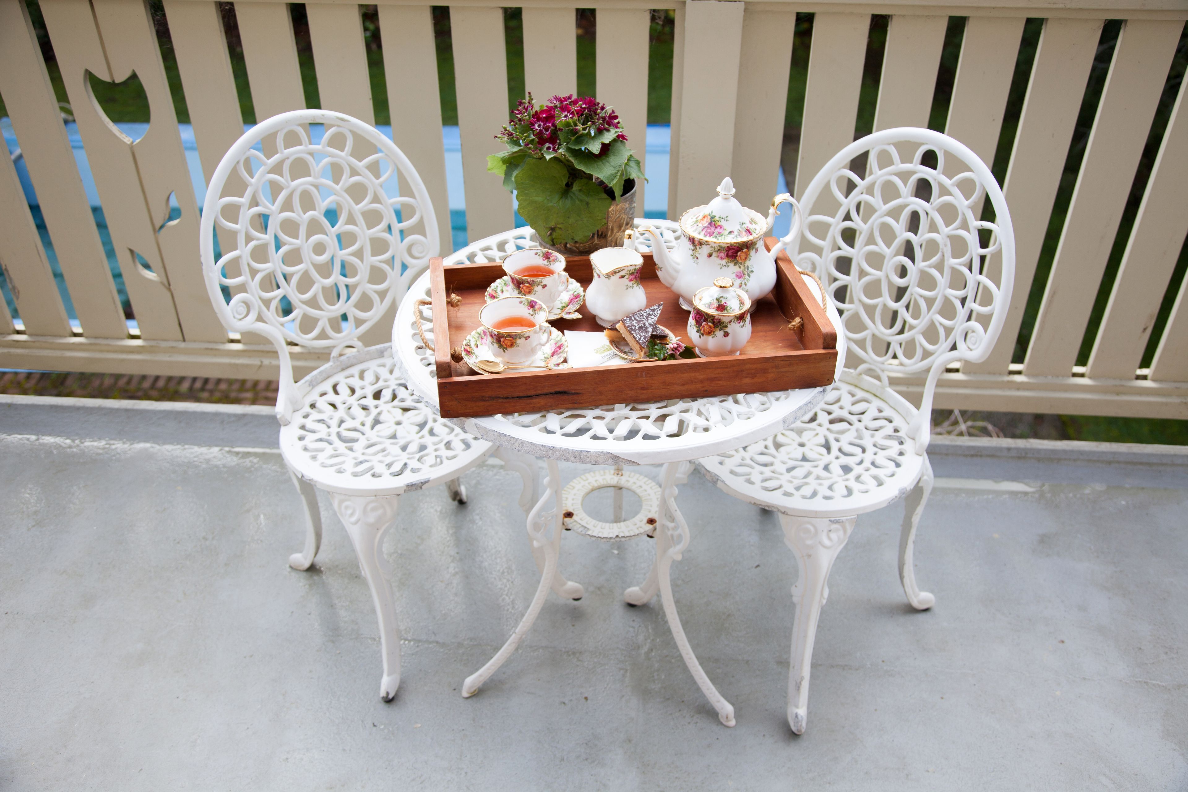 Two ornate white chairs with a white table between them, holding a tray with tea cups, teapot, sugar bowl, and a flower vase.