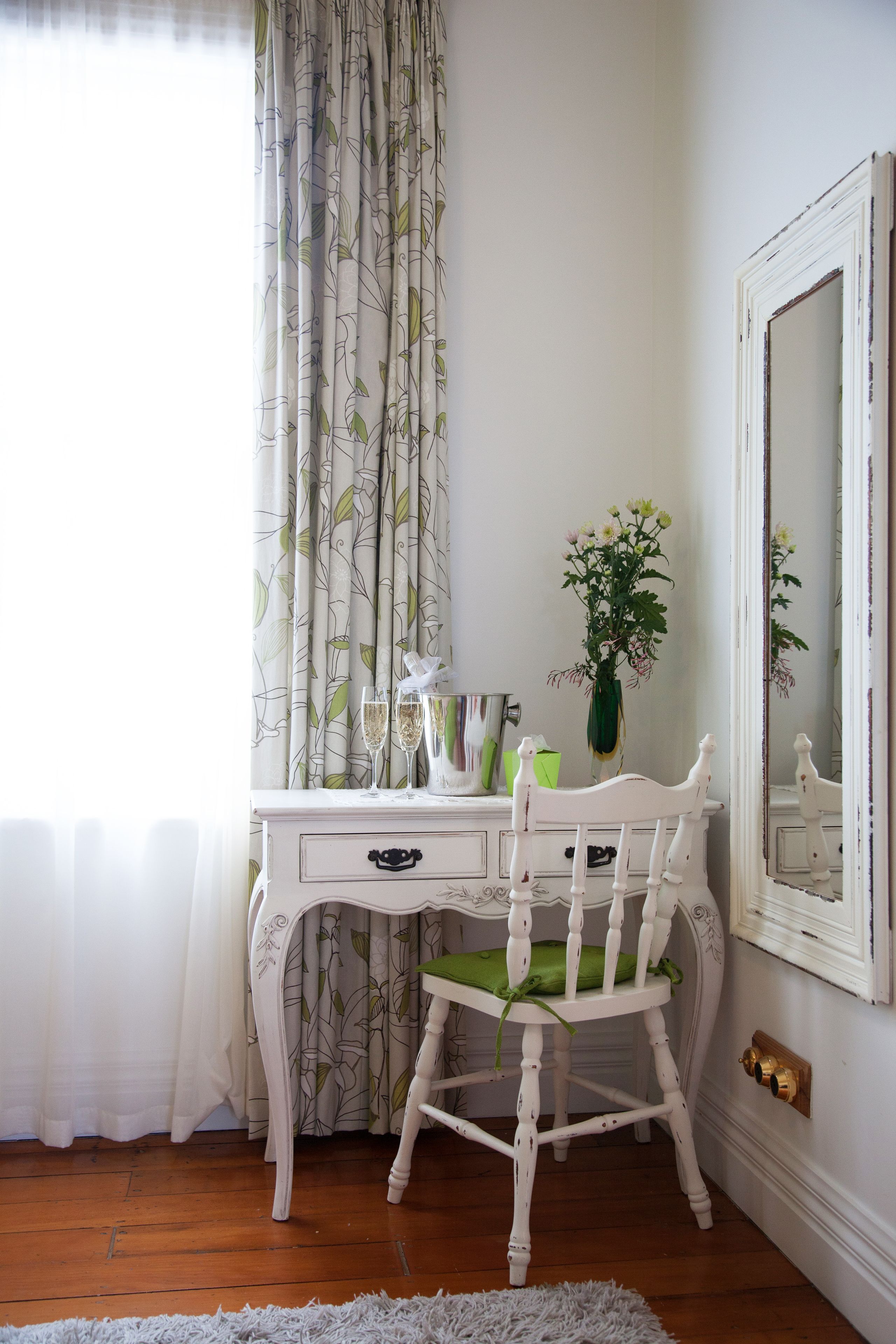 A white wooden desk with drawers, a green cushioned chair, a vase with flowers, and a mirror on the wall.