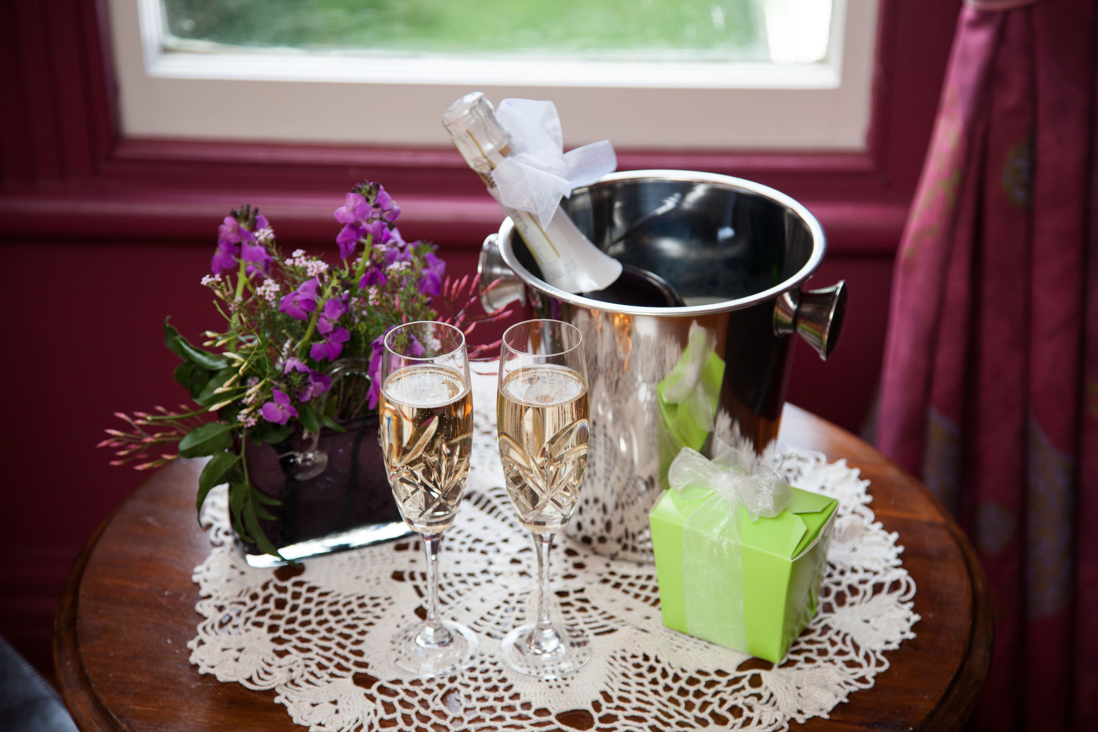 A table with a metal bucket containing a bottle, two glasses of champagne, a green gift box, and a potted plant with purple flowers.