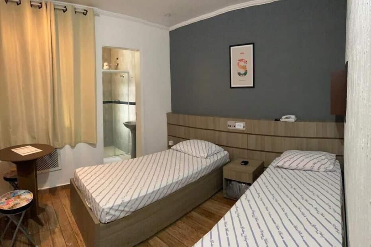 A small, compact room with two single beds, a small table, a chair, a mirror, and a shower area.