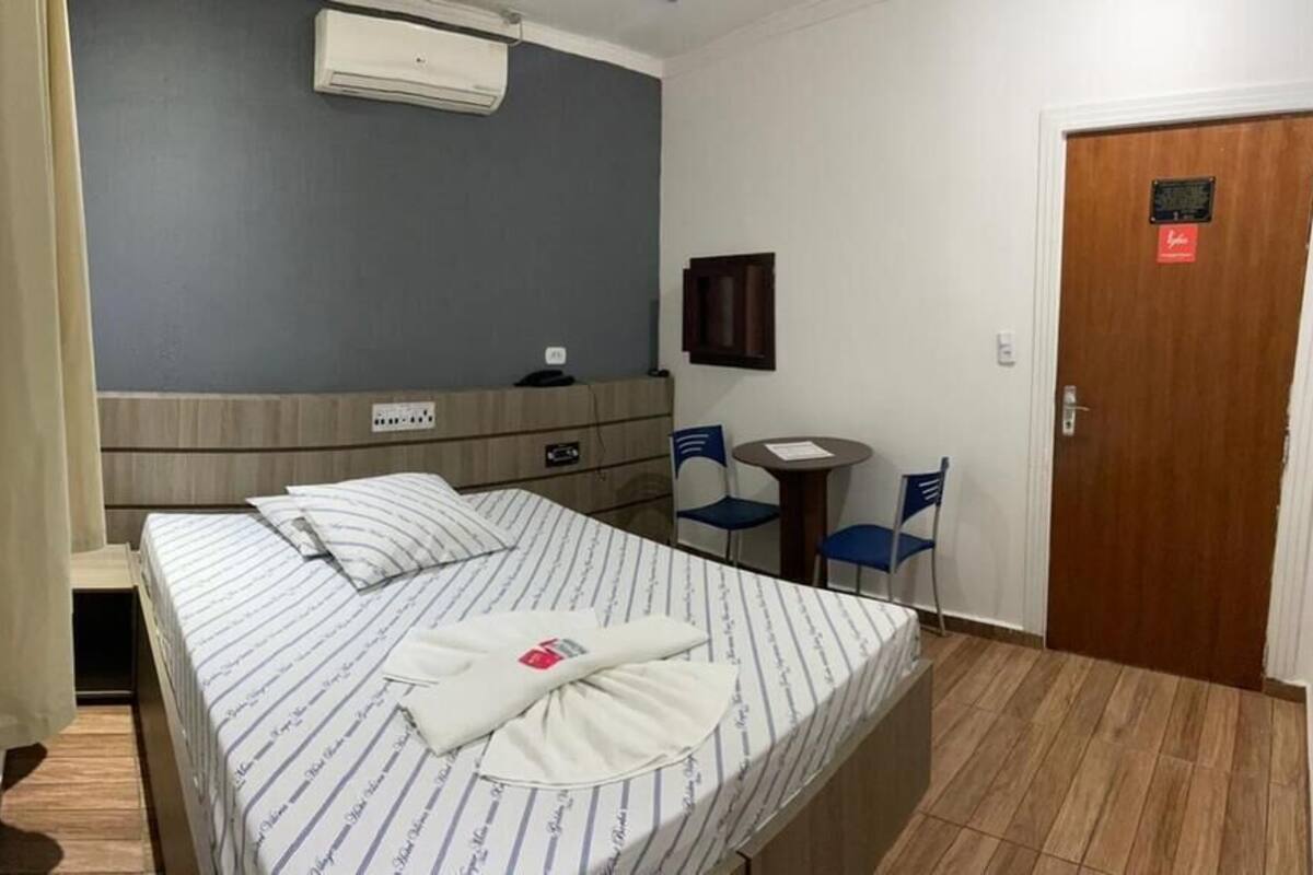 A hotel room with a bed, a desk, a chair, a door, and an air conditioning unit.