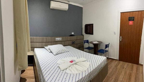 A hotel room with a bed, a desk, a chair, a door, and an air conditioning unit.