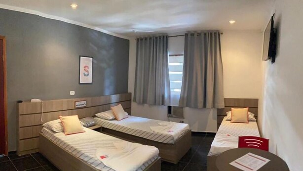 A hotel room with two single beds, a small table, a TV, and a window with curtains. - Hotel Belém (São Paulo)