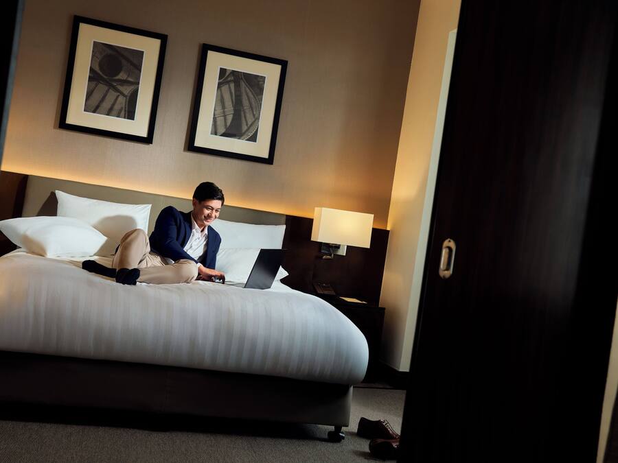 A hotel room with a large bed, a desk with a laptop, two framed pictures on the wall, and a nightstand with a lamp.