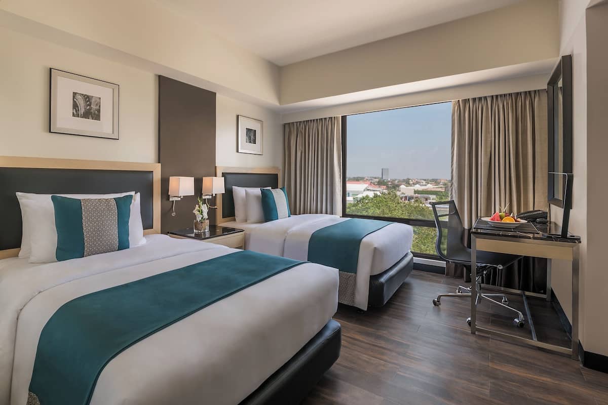 A hotel room with two beds, a desk, a chair, and a large window offering a city view.