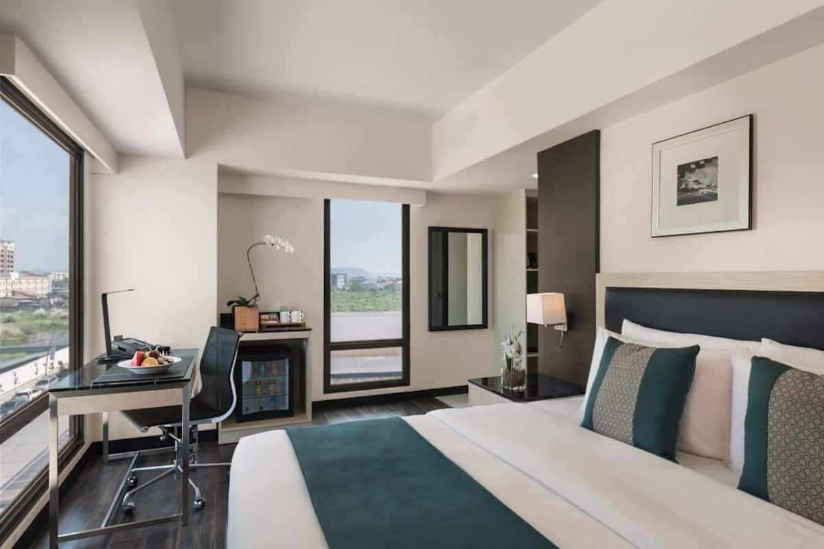 A hotel room with a large bed, a desk with a chair, a small table with a lamp, and a view of the city.