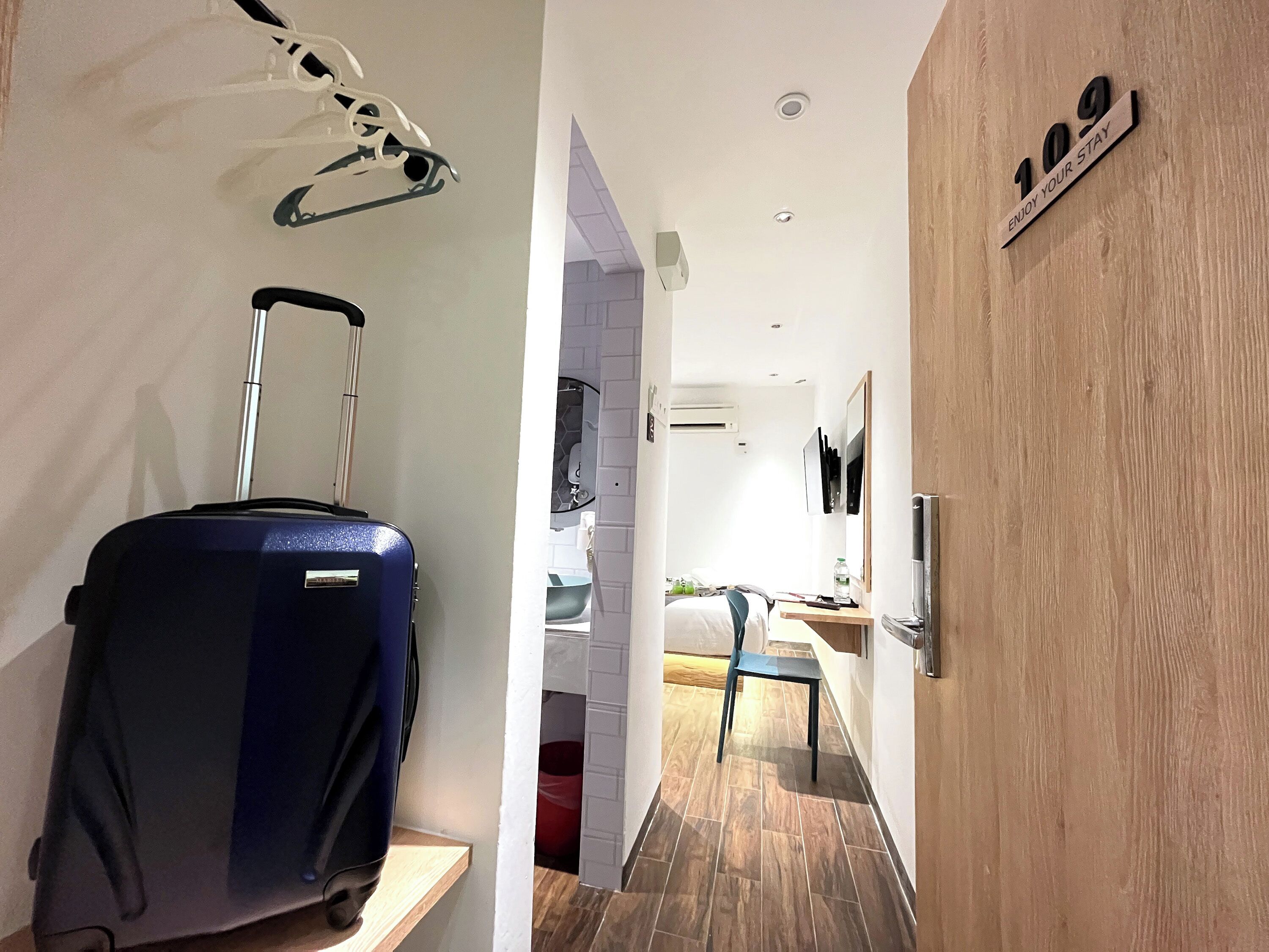 Habitación de hotel con puerta de madera, cartel que dice '109 DISFRUTA TU ESTANCIA', una maleta azul y una cama con almohada roja.
