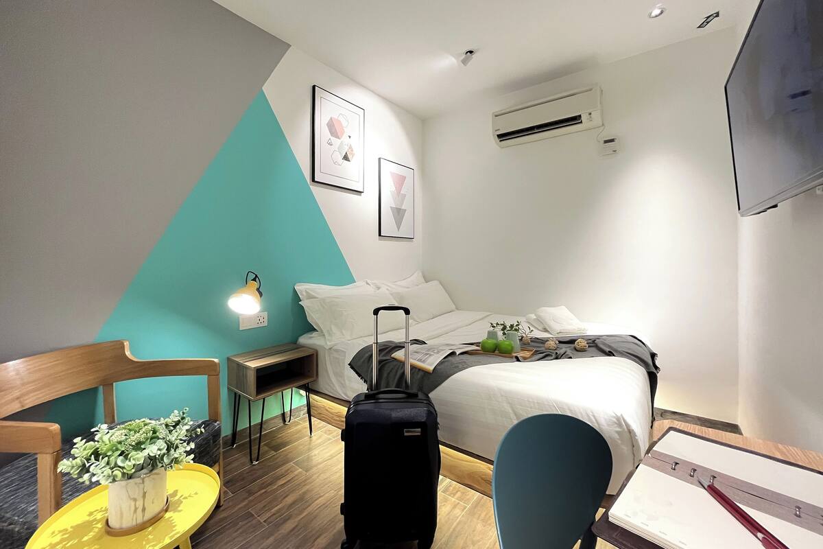 A hotel room with a bed, a desk, a chair, a suitcase, a plant, and a TV.