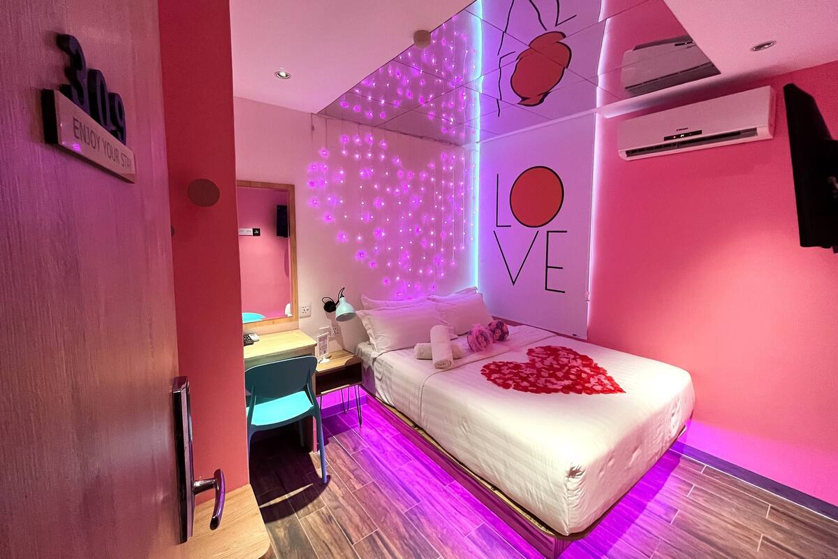 A modern bedroom with a bed, a desk, and a wall with 'LOVE' and 'ENJOY YOUR STAY' signs.