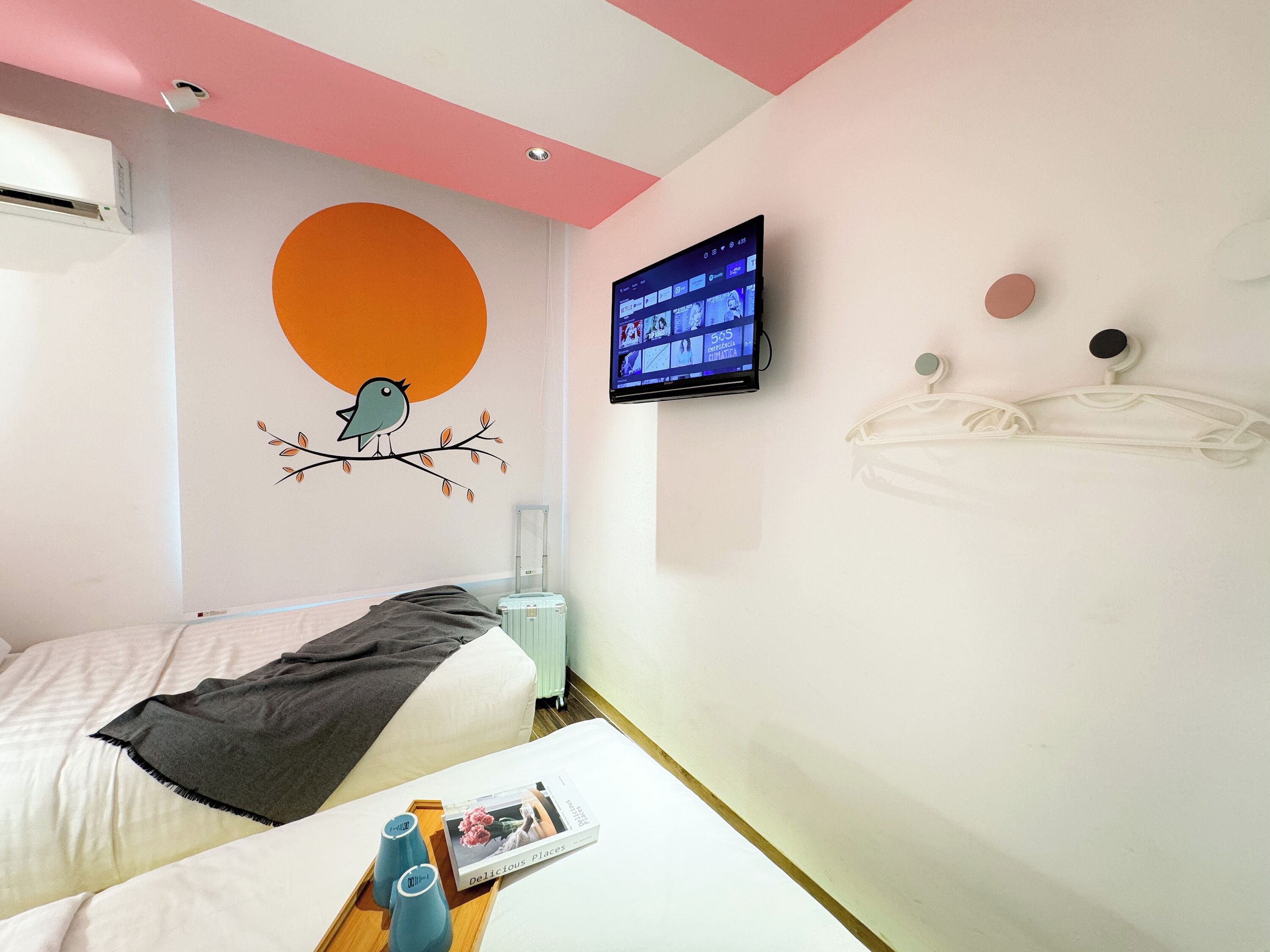 A hotel room with a bed, a wall-mounted TV, a wall decoration, and a bedside table with a magazine and bottles.
