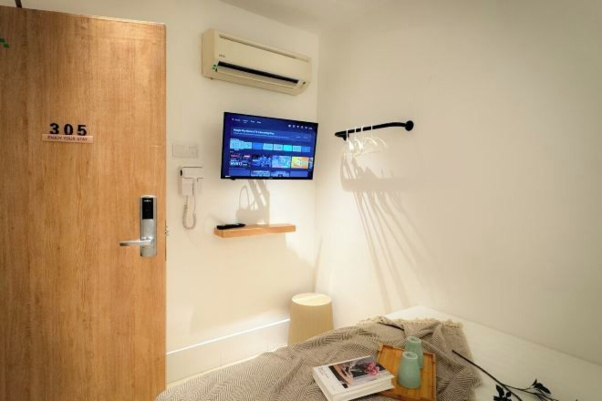 A hotel room with a wooden door marked '305', a mounted TV, a bedside lamp, a bed with a patterned blanket, and a small wooden side table with a book and a vase.