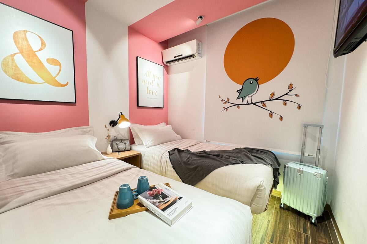 Habitación de hotel con dos camas, una mesita de noche con lámpara, una bandeja con un libro y una pared con un mural de aves.