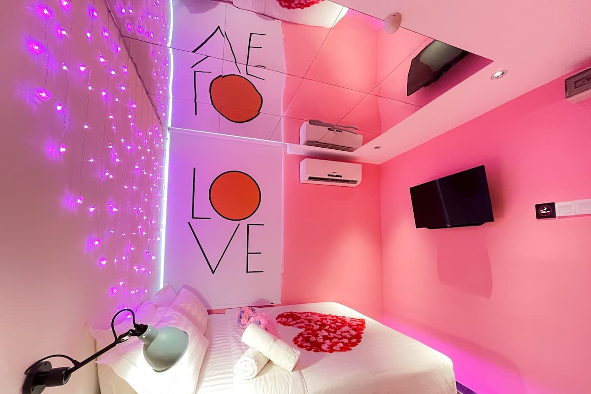 Un dormitorio moderno con pared rosa, cama con ropa de cama blanca, lámpara de noche y televisor montado en la pared.