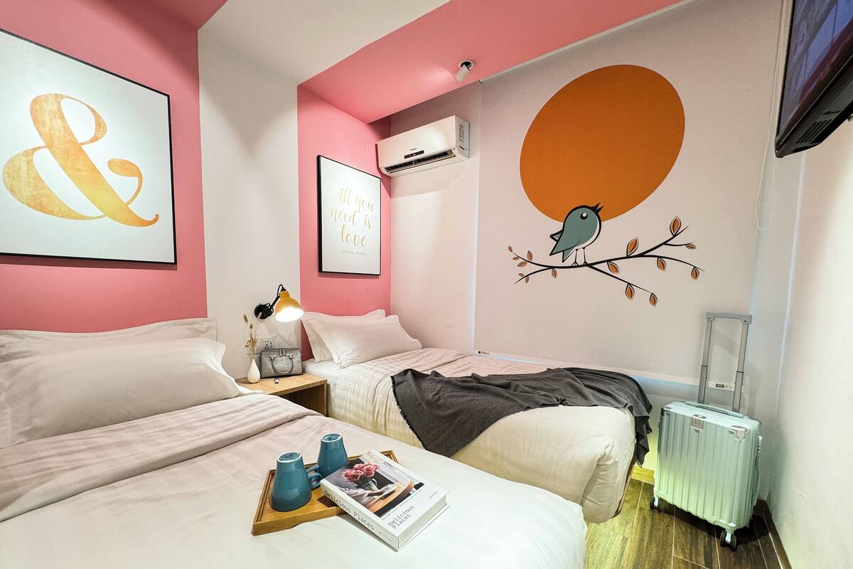 A hotel room with two beds, a bedside table with a lamp, a tray with a book, and a wall with a bird mural.