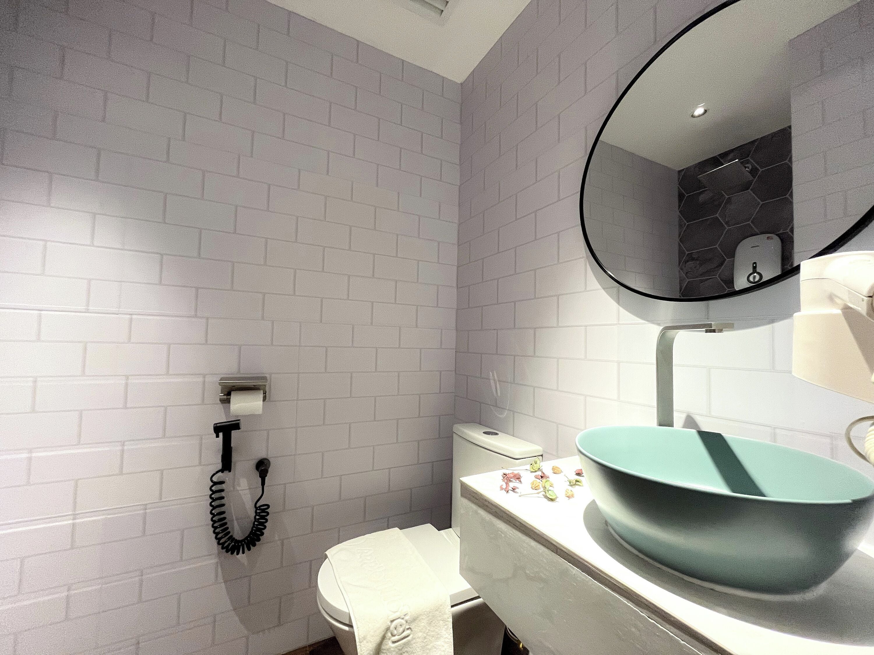 Une salle de bain avec des murs recouverts de carreaux blancs, des toilettes, un lavabo doté d’un grand bac et un miroir avec un pare-éclaboussures en forme d’hexagone.