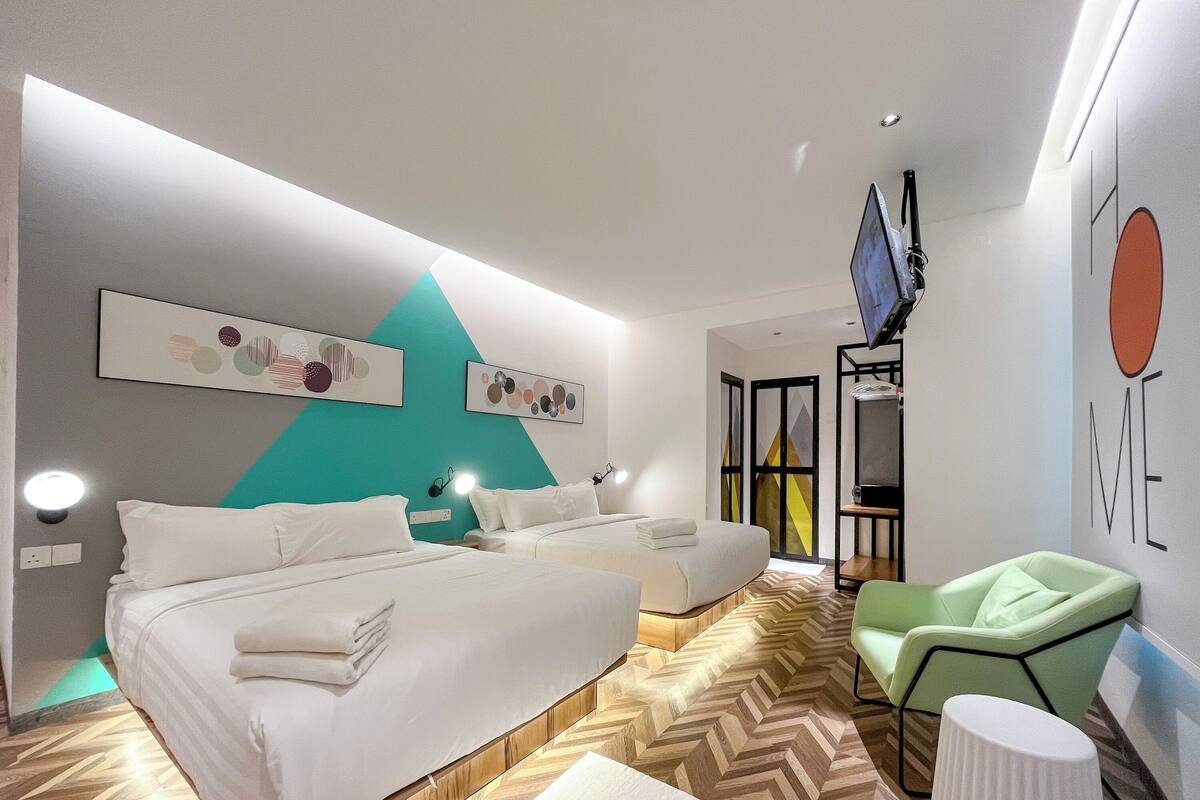 A modern hotel room with a large bed, two smaller beds, a green armchair, a small table, and artwork on the walls.