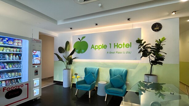 A hotel reception area with a vending machine, a large green apple sign, and two blue chairs. - Apple 1 Hotel Queensbay (George Town)