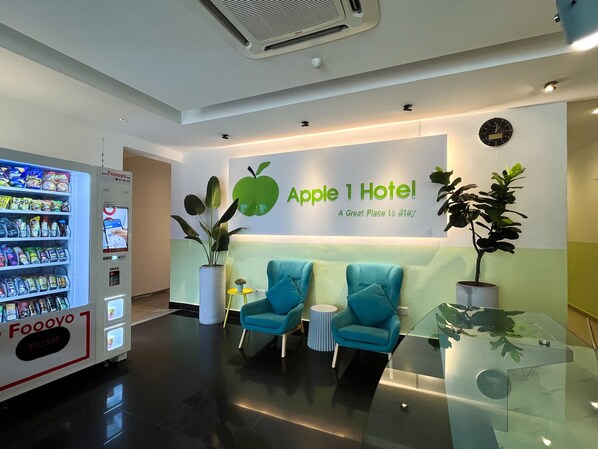 A hotel reception area with a vending machine, a large green apple sign, and two blue chairs. - Apple 1 Hotel Queensbay (George Town)