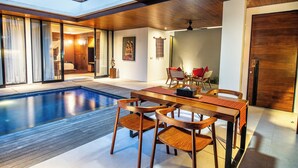 A modern indoor pool area with wooden flooring, a dining table with chairs, and a ceiling fan. - Abia Villa and Suite Legian (Legian)