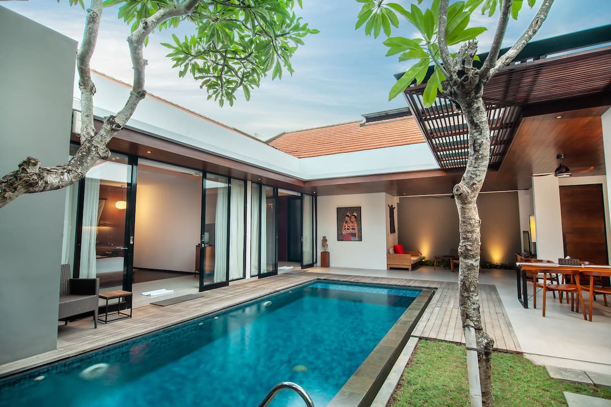 A modern outdoor pool area with a wooden deck, a glass-enclosed room, and a dining area with a table and chairs.