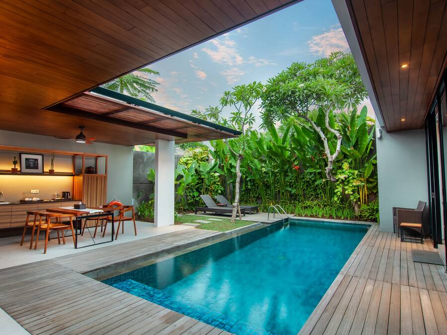 Un espace piscine extérieur moderne, doté d’une terrasse en bois, d’un ensemble de mobilier de jardin et d’un coin cuisine offrant une vue sur une végétation luxuriante.