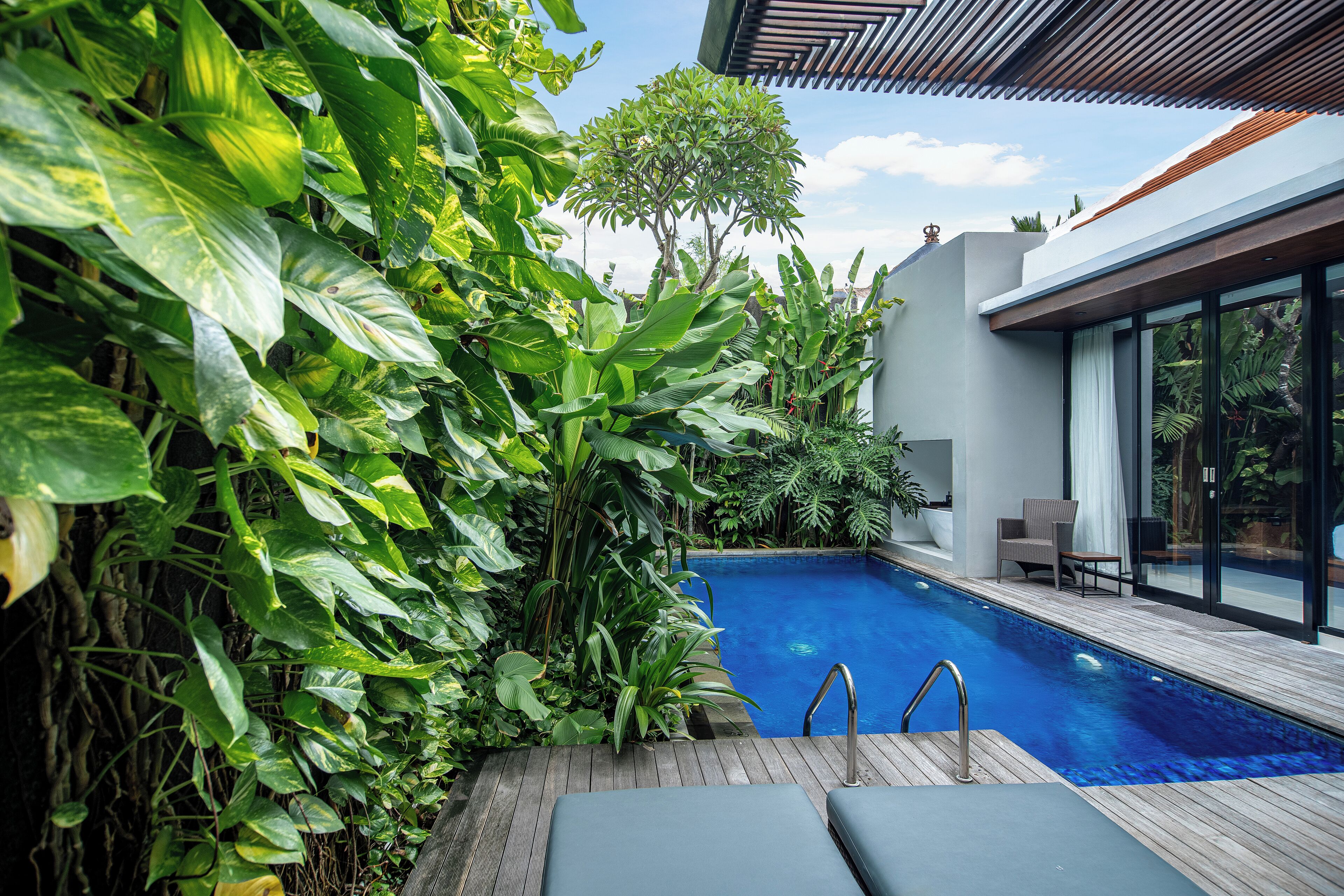 A modern outdoor pool area with lush greenery, a wooden deck, and a seating area with a view of the pool.
